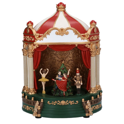 A decorative music box theatre featuring a red and white striped dome, gold trim, red curtains, and small figurines including a ballerina, a Christmas tree, and a nutcracker on a green base against a white background.