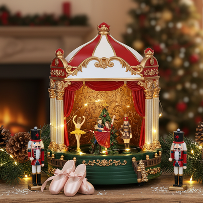 A decorative holiday music box or miniature stage set on a tabletop, featuring a red-and-white striped dome, gold trim, red curtains, small figurines, nutcracker figures, pink ballet slippers, and warm string lights. Christmas trees and a softly blurred indoor holiday background appear behind it.