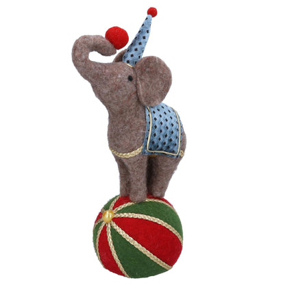 A felt elephant toy balancing on a red and green striped ball, wearing a blue polka-dot pointed hat and a blue patterned saddle with gold trim, isolated on a white background.