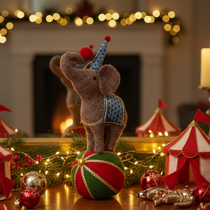 A festive felt elephant decoration wearing a blue party hat and patterned saddle, standing on a red and green striped ball in a decorated holiday scene with lights, ornaments, and small circus tents.