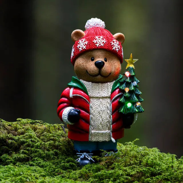 A decorative teddy bear wearing a Christmas hat and holding a miniature Christmas tree, placed on a mossy surface.