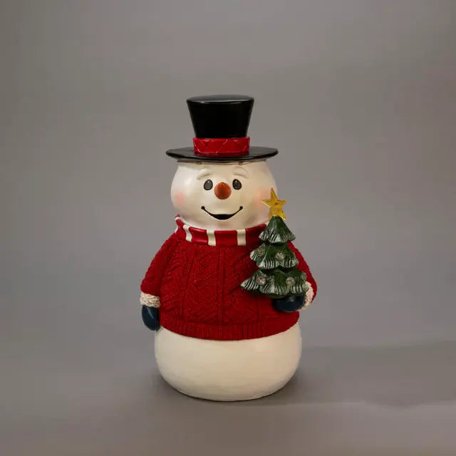 Decorative snowman figurine with red top hat and sweater holding a small Christmas tree on a gray background.