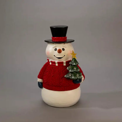 Decorative snowman figurine with red top hat and sweater holding a small Christmas tree on a gray background.