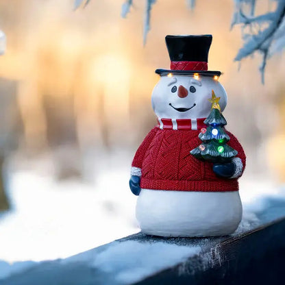 A decorative snowman figure with multicolor and warm white LED lights, dressed in a red sweater and a black hat, holding a small Christmas tree, placed outdoors during winter.