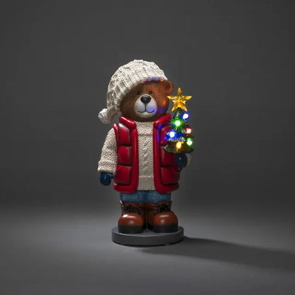 A decorative teddy bear wearing a knitted hat and a red and white coat, holding a Christmas tree with multicolored LED lights.