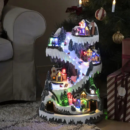 A mechanical ski slope Christmas decoration featuring multicolored LED lights, with houses and a tree on a tiered platform, placed on a floor next to a chair and a wrapped gift box.