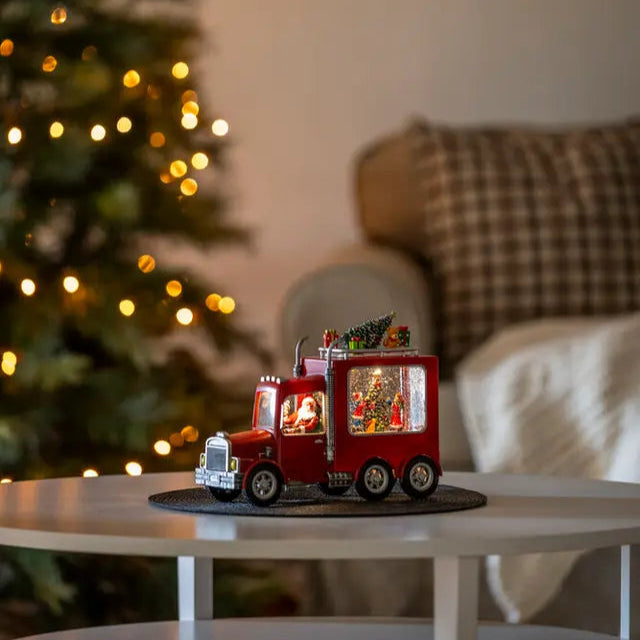 A decorative Santa Truck with LED lights, placed on a table in front of a Christmas tree, showcasing a miniature festive scene.