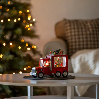 A decorative Santa Truck with LED lights, placed on a table in front of a Christmas tree, showcasing a miniature festive scene.