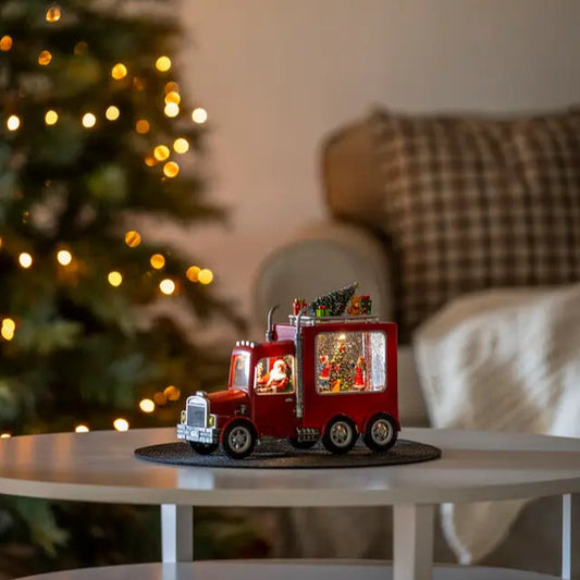 A decorative Santa Truck with LED lights, placed on a table in front of a Christmas tree, showcasing a miniature festive scene.