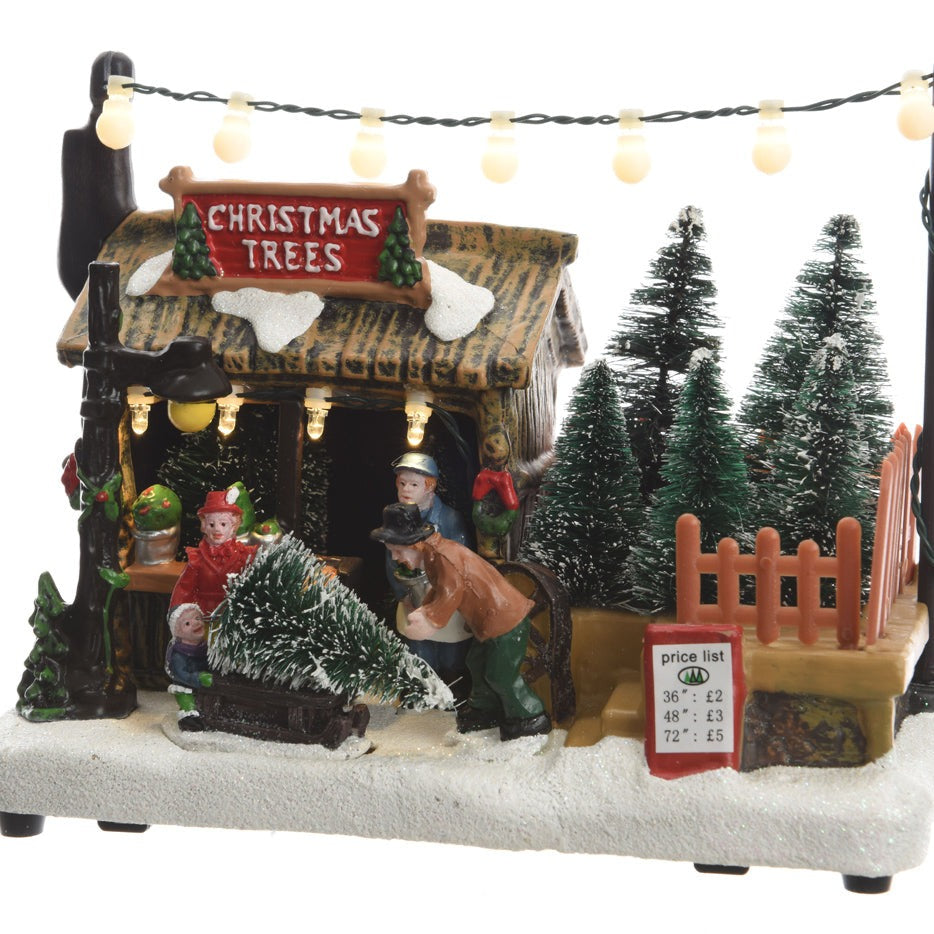 A festive Christmas village scene decoration featuring a tree seller with a lit storefront, surrounded by Christmas trees and warm white lights.