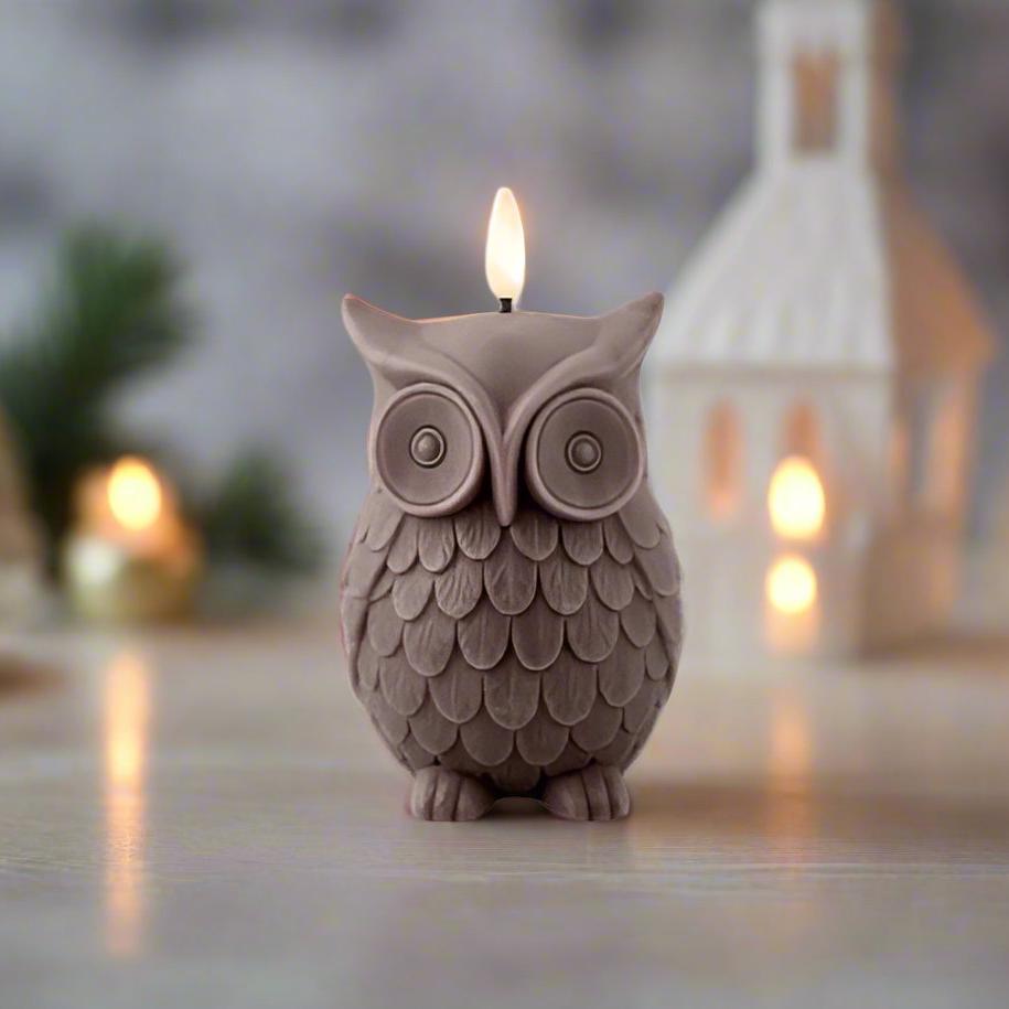 A brown wax candle in the shape of an owl, with a lit wick, placed on a table with a blurred warm light background featuring a Christmas tree and house.