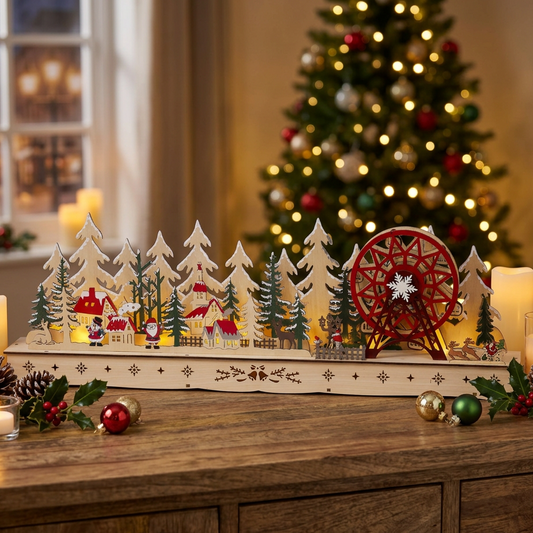 A wooden Christmas village tabletop decoration with a winter forest scene, small figures, evergreen trees, a red Ferris wheel, and a sleigh, displayed on a wooden surface in front of a softly blurred decorated Christmas tree.