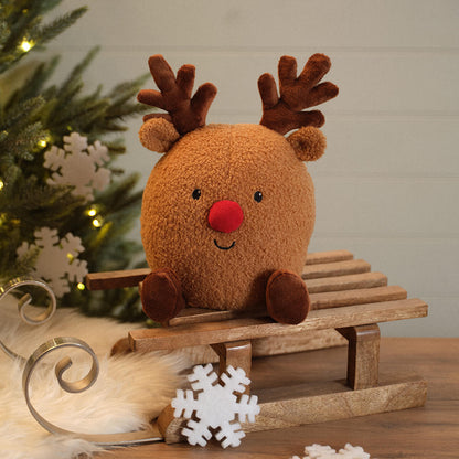 A round brown plush reindeer door stop  with a red nose and dark brown antlers sits on a small wooden sled. The scene is styled with a Christmas tree, white snowflake decorations, and a warm holiday background.