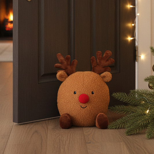 A round brown plush reindeer door stopper with a red nose and embroidered face sits on a wooden floor in front of an open dark door, with Christmas tree branches and warm string lights visible around it.