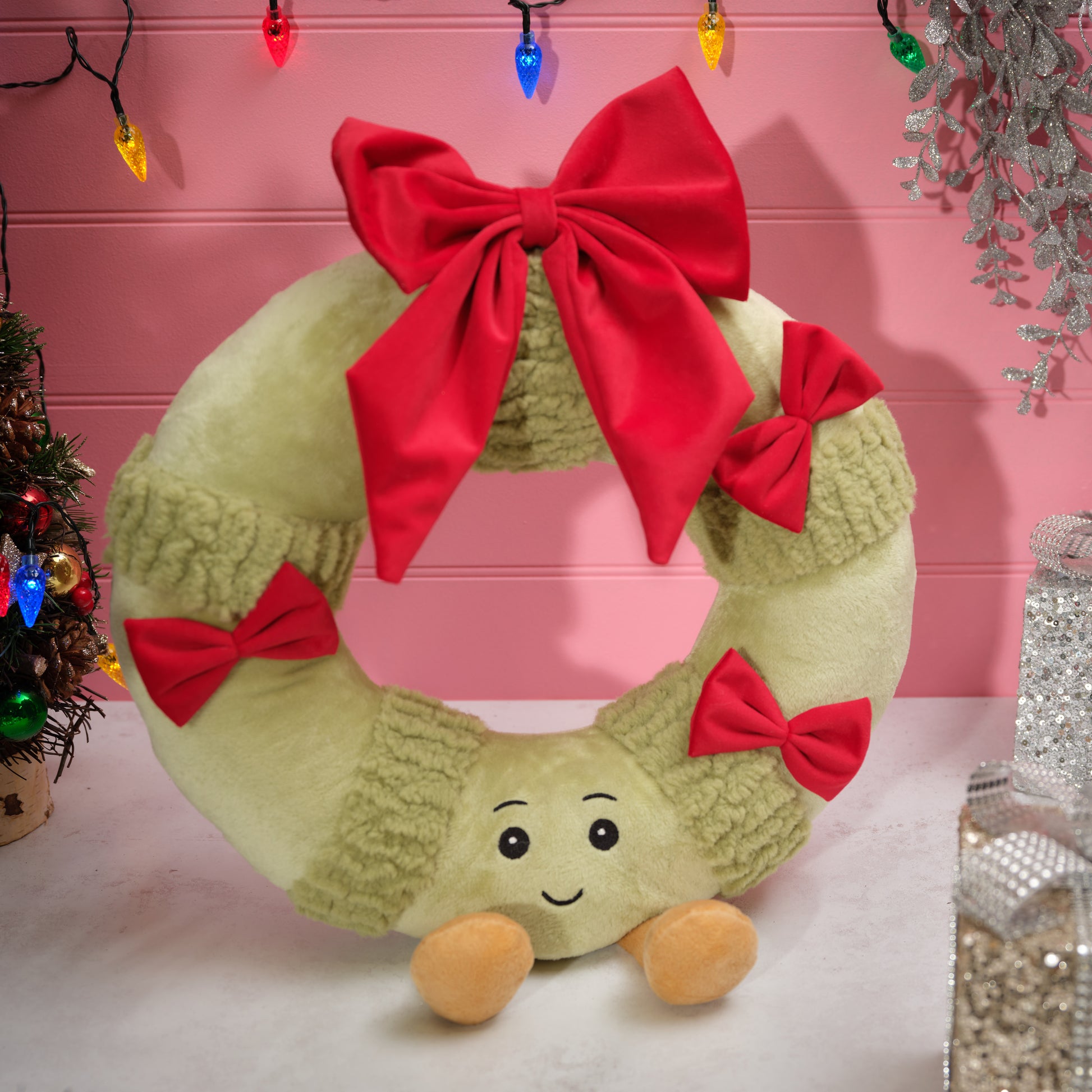 A plush Christmas wreath decoration in light olive green with textured knit-like bands, large red bows, and a smiling face with small tan feet, displayed against a pink holiday-themed backdrop.