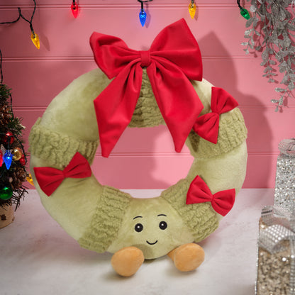 A plush Christmas wreath decoration in light olive green with textured knit-like bands, large red bows, and a smiling face with small tan feet, displayed against a pink holiday-themed backdrop.