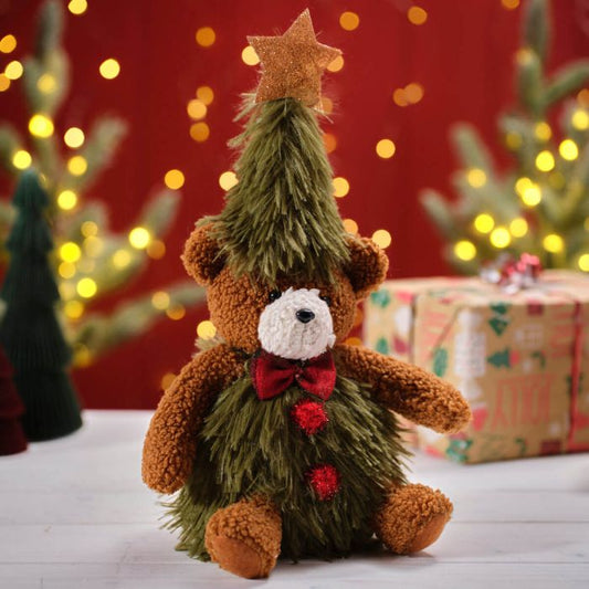 A plush sitting bear with a Christmas tree on its back and a star on top, adorned with red ribbon and a red bow, against a festive background with bokeh lights and gift boxes.