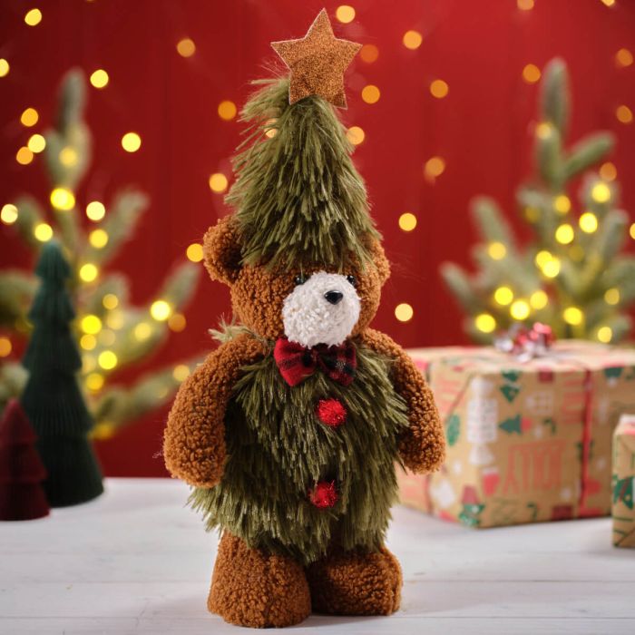 A plush toy bear with a Christmas tree design, featuring a star on its head and decorated with red and green festive decorations, standing against a holiday-themed backdrop with bokeh lights and gift boxes.