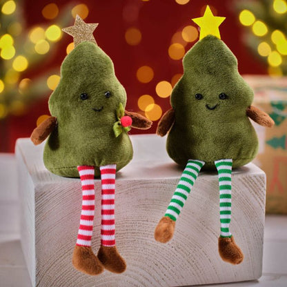 Two plush Christmas tree shelf sitters with one wearing a red star and the other a green star, seated on a wooden block with bokeh lights in the background.