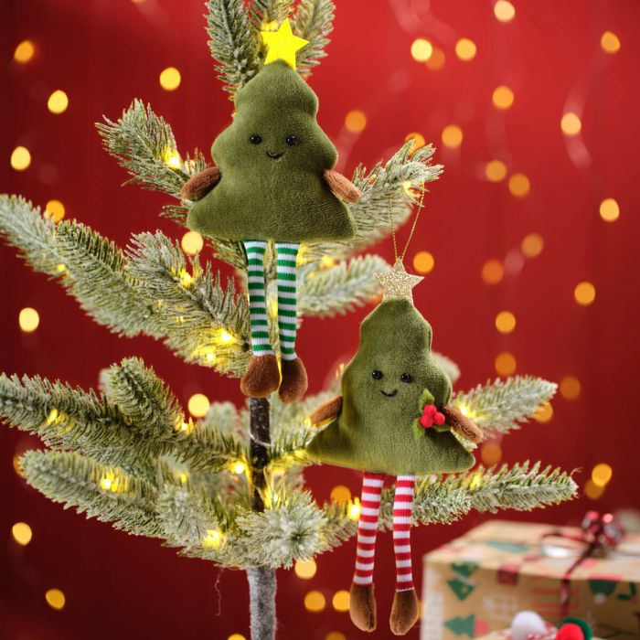 Two Christmas tree hangers, one green tree-shaped and one with a face wearing a striped scarf, hanging on a Christmas tree with lights in the background.