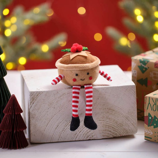 A fabric shelf sitter resembling a mince pie with legs and a festive design, placed on a wooden block with Christmas decorations in the background.
