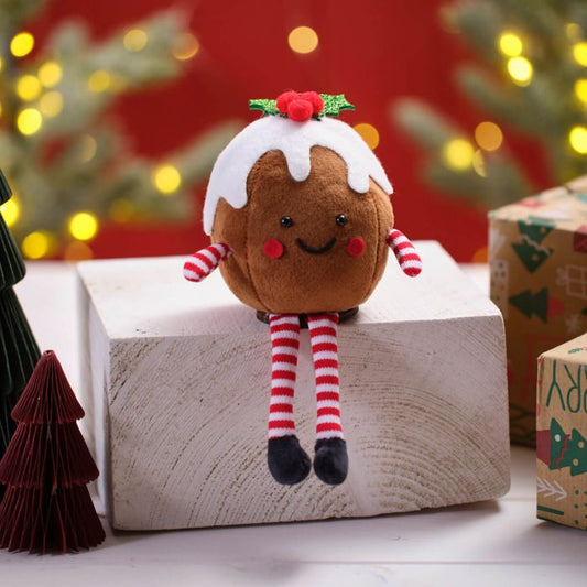 A fabric shelf sitter shaped like a Christmas pudding, with a red and white striped design on its legs and a smiley face on its top. It is placed on a wooden surface with festive decorations in the background.