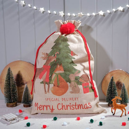 A beige Christmas sack with red trim and festive decorations, including a Christmas tree and reindeer, displayed on a wooden background with Christmas lights and small ornaments.