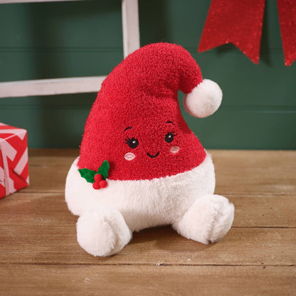 A plush Christmas Santa hat toy with a red fuzzy hat, white fluffy body and cuff, embroidered face, and a small holly accent, sitting on a wooden surface.