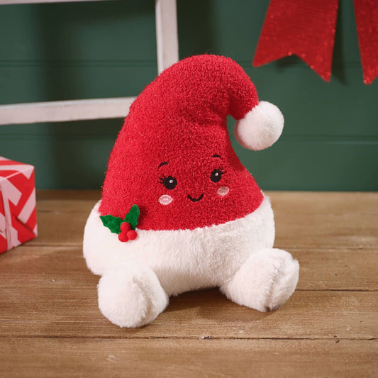 A plush Christmas Santa hat toy with a red fuzzy hat, white fluffy body and cuff, embroidered face, and a small holly accent, sitting on a wooden surface.