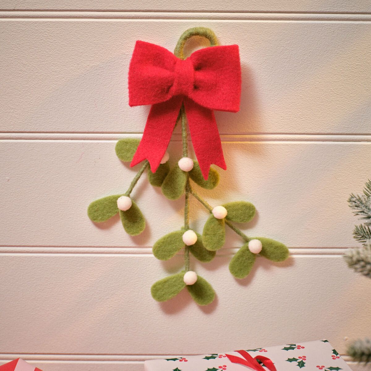 A felt mistletoe holiday decoration with a large red bow, green leaves, and small white berries hanging against a white paneled wall.