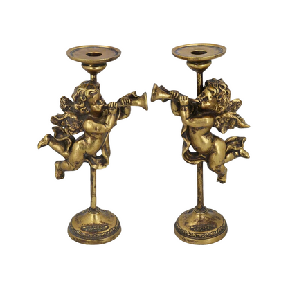 Two gold-colored candlesticks featuring cherub figures with trumpets, placed on a wooden surface with pine branches in the background.