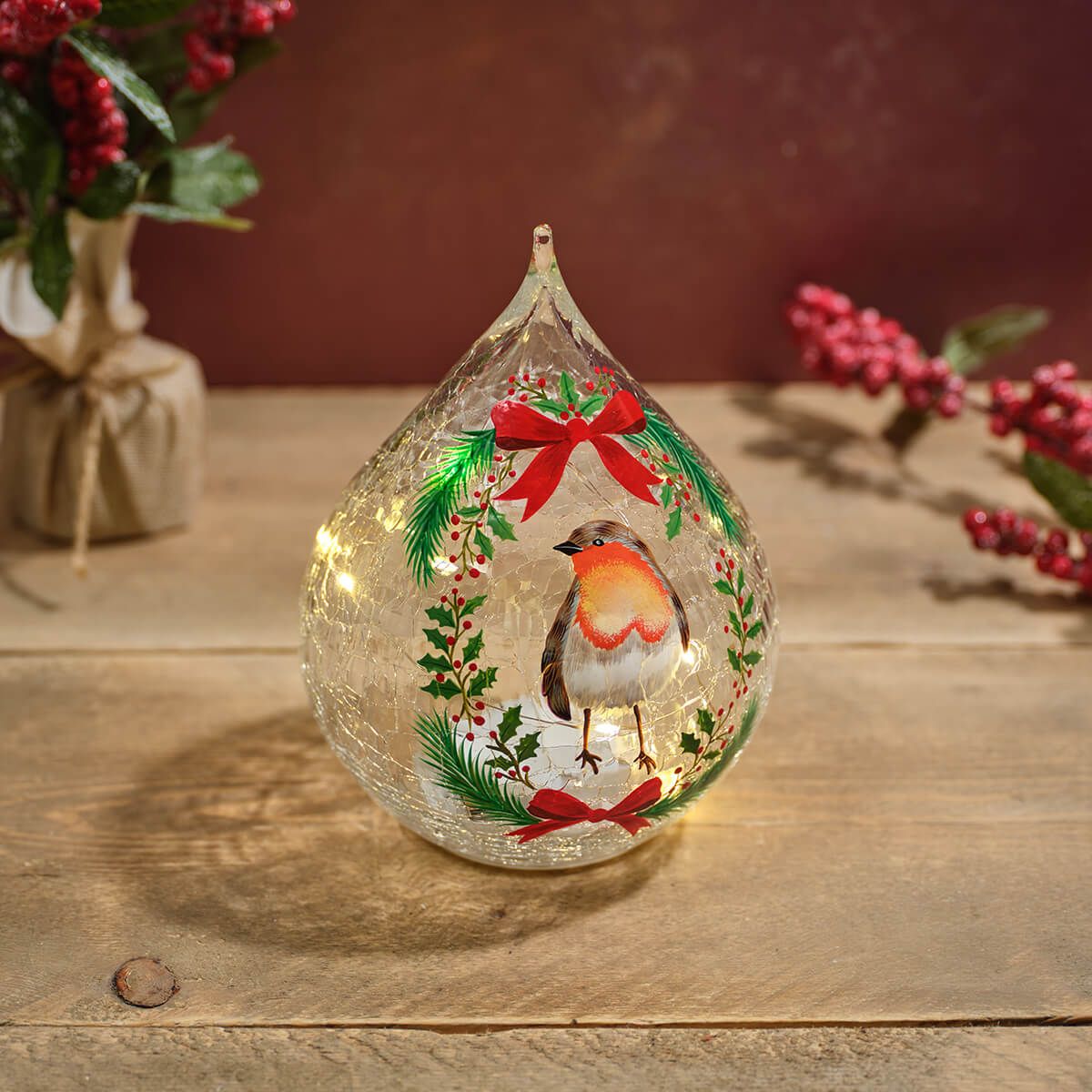 A teardrop-shaped clear glass ornament with a robin bird and Christmas wreath design, placed on a wooden surface with warm fairy lights inside.