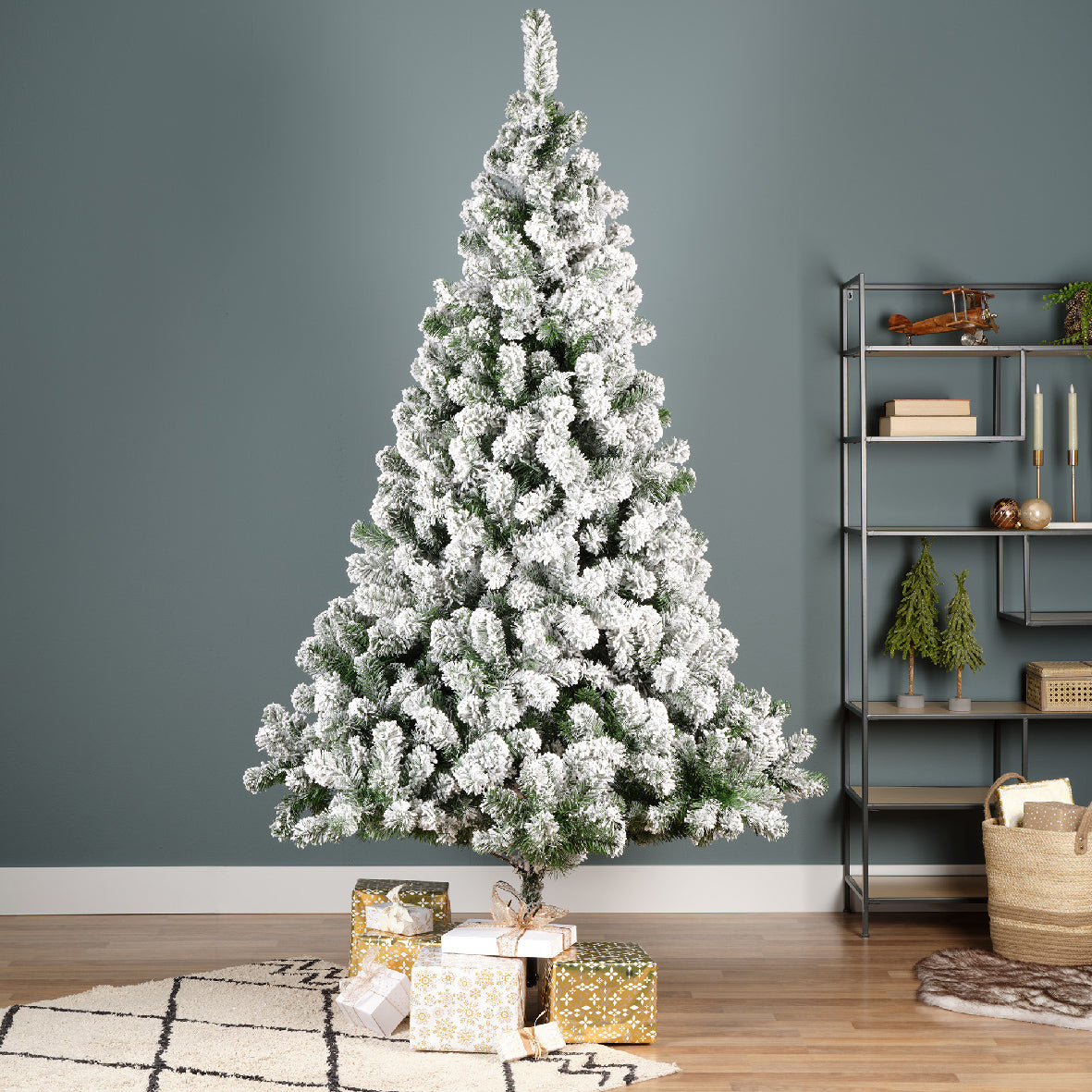 A 7ft tall artificial snowy green Christmas tree with metal base, placed in a home setting, surrounded by holiday decorations and gifts.