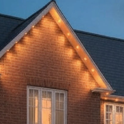 Warm white outdoor string lights illuminated along the gable and roofline of a brick house at dusk.