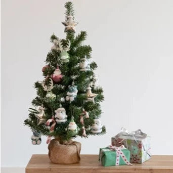 A pre-decorated mini Christmas tree with a green trunk and mixed color baubles, placed on a wooden surface with a gift box and decorative star ornaments nearby.