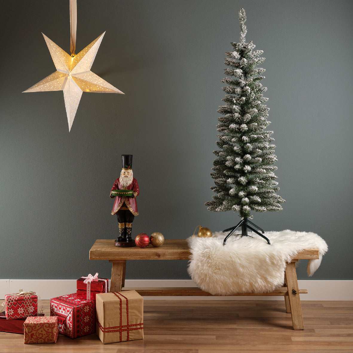 Artificial Christmas tree with snowy pencil pine branches and a classic profile, placed on a wooden bench with presents and a decorative star on a green wall in the background.