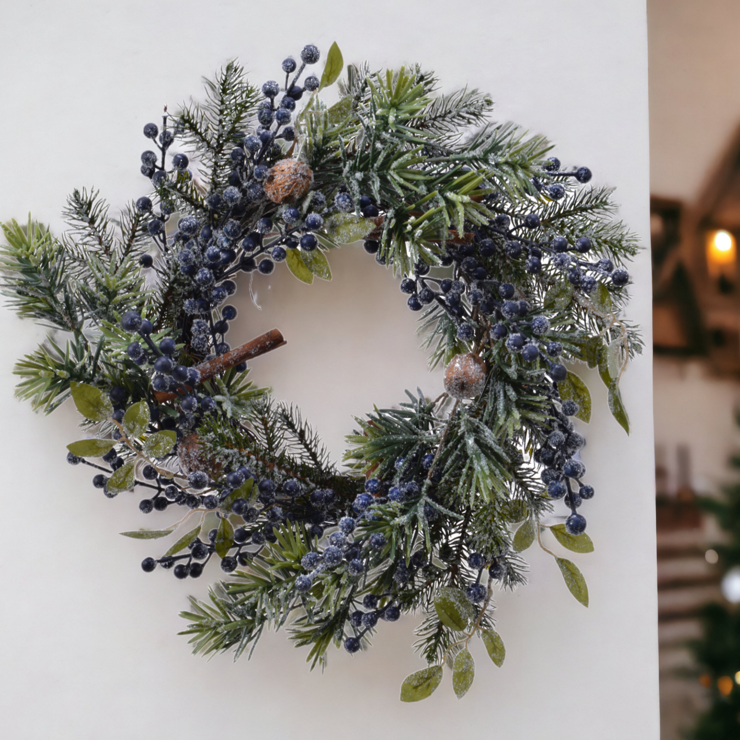 A round Christmas wreath decorated with frosted blue berries, green foliage, cinnamon sticks, and nuts, displayed against a neutral background.