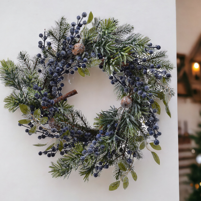 A round Christmas wreath decorated with frosted blue berries, green foliage, cinnamon sticks, and nuts, displayed against a neutral background.