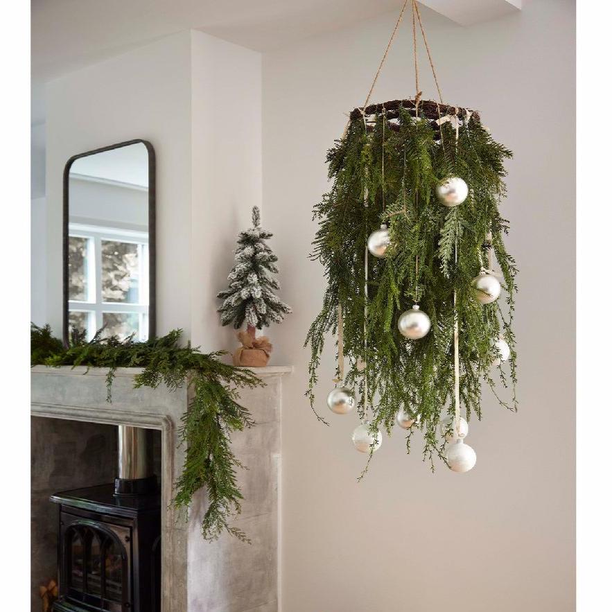 A hanging Christmas wreath with mixed green foliage and white ornamental balls, displayed indoors near a fireplace.