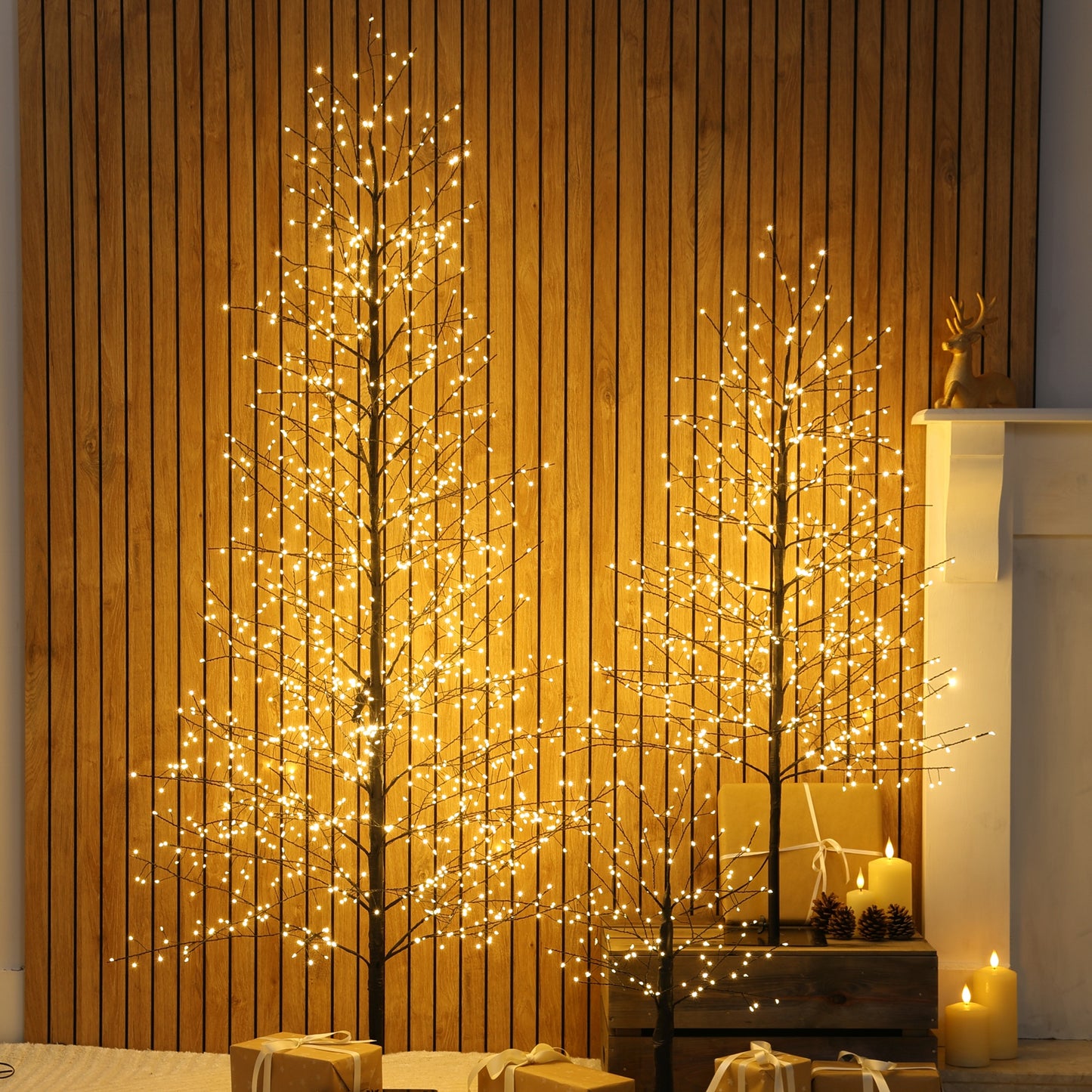 Two tall artificial Christmas twig trees with warm white LED lights, displayed in a cosy room setting with a wood-panelled wall, candles, and wrapped gifts.