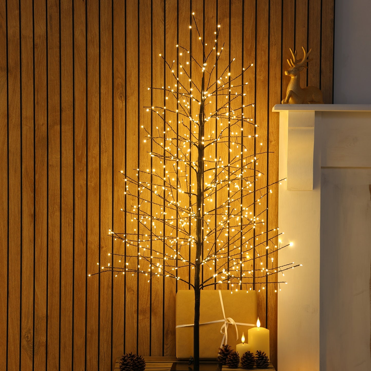 A decorative black twig tree with warm white LED string lights, styled indoors against a wood-panelled wall beside a white mantel with a small deer figurine and candles.