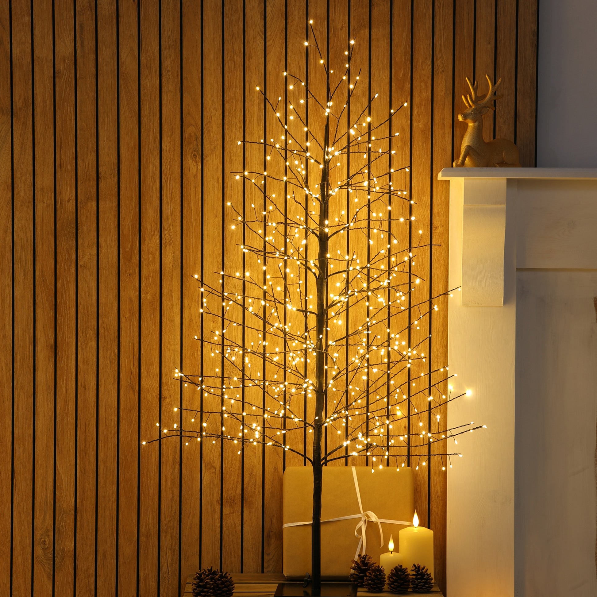 A decorative black twig tree with warm white LED string lights, styled indoors against a wood-panelled wall beside a white mantel with a small deer figurine and candles.