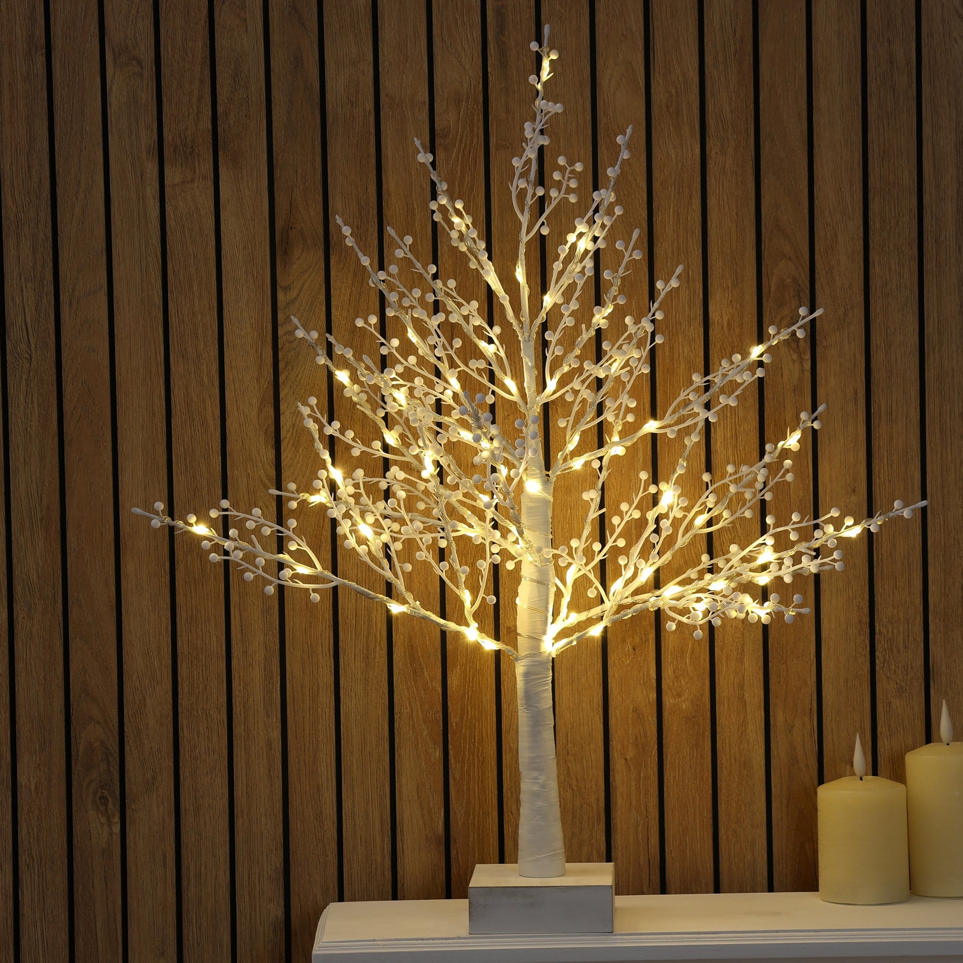 A decorative illuminated tree with a white trunk and branching limbs covered in warm white fairy lights, displayed on a white surface against a dark wood slat wall, with two lit candles at the side.
