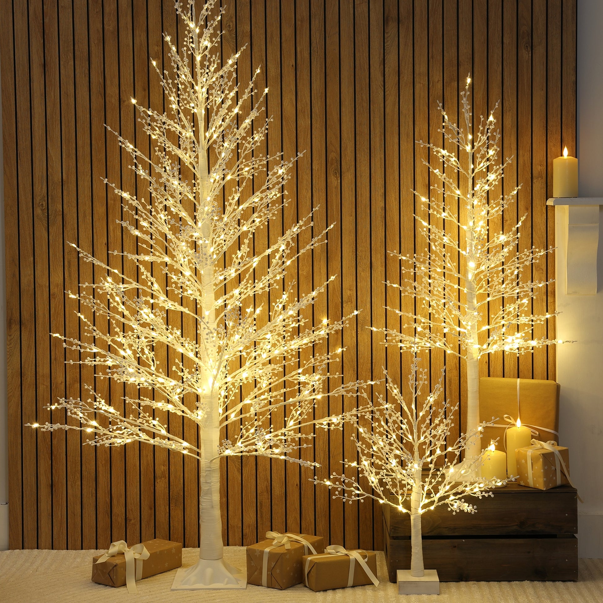 Three illuminated LED Christmas twig trees with berry lights with warm white glow, displayed in different sizes against a vertical wood-panelled wall with wrapped gifts at the base.
