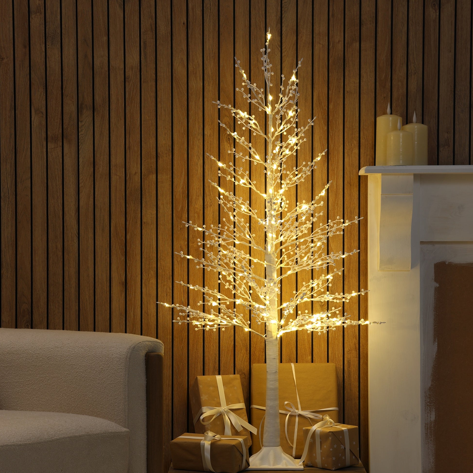 A decorative artificial twig tree with glowing warm white string lights stands indoors against a wood-paneled wall, with wrapped gift boxes at its base and part of a fireplace and sofa visible.