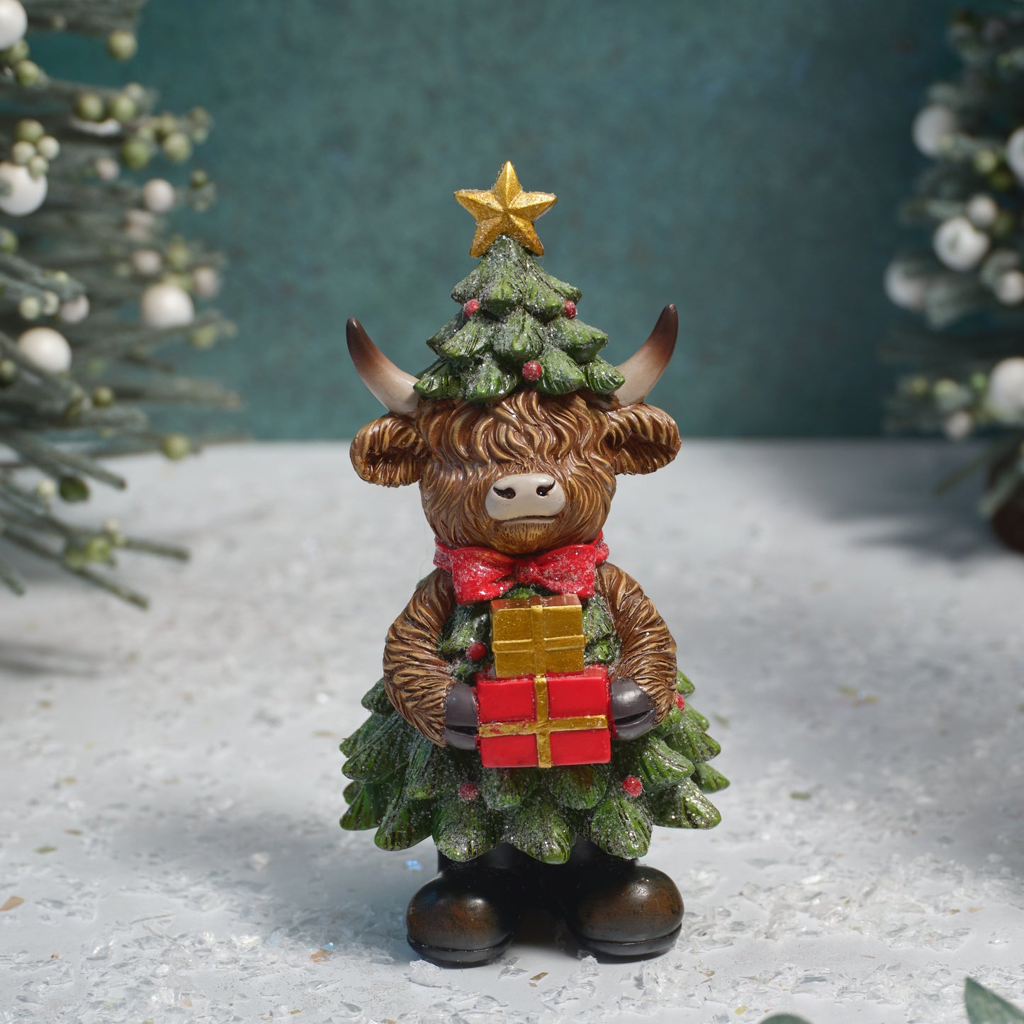 A decorative Highland cow figurine dressed as a Christmas tree, holding a red and gold gift box, set against a snowy, festive backdrop.