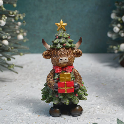 A decorative Highland cow figurine dressed as a Christmas tree, holding a red and gold gift box, set against a snowy, festive backdrop.