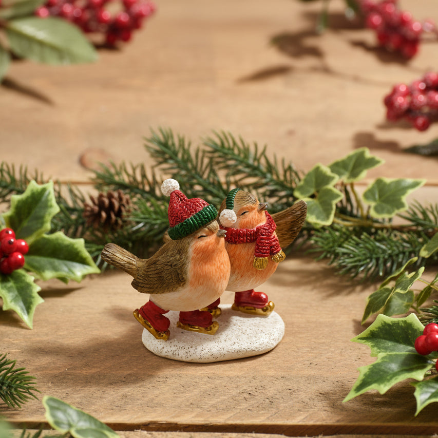 Two small robin figurines wearing festive red and green holiday hats and scarves, standing on a white base on a wooden surface.