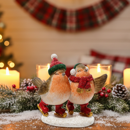 Two small Christmas robin figurines posed together on a round base, wearing red scarves and hats with gold accents, set in a festive holiday scene with candles, pinecones, and blurred lights in the background.