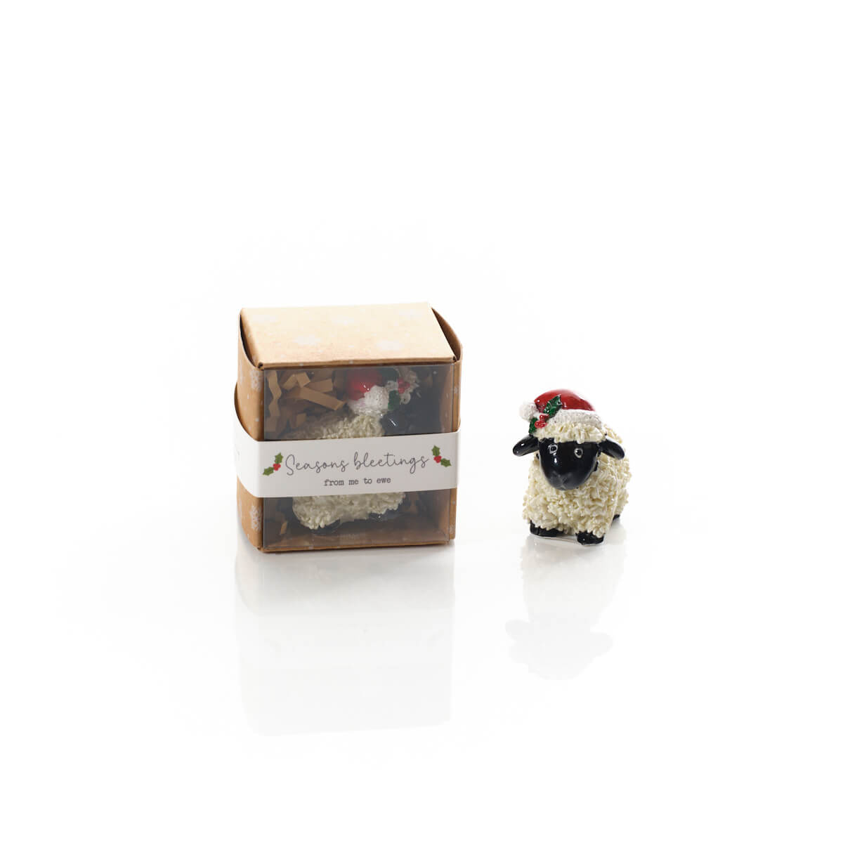 A small packaged ornament or figurine shown next to its box on a white reflective surface. The product appears to be a fluffy white sheep with a black face and a red Santa hat, with a paper band on the box reading 'Season's bleetings from me to ewe'.